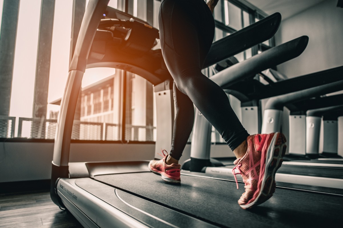 A person running on a treadmill, wearing pink sneakers and black leggings, in a sunlit gym with large windows. The tone is energetic and focused.