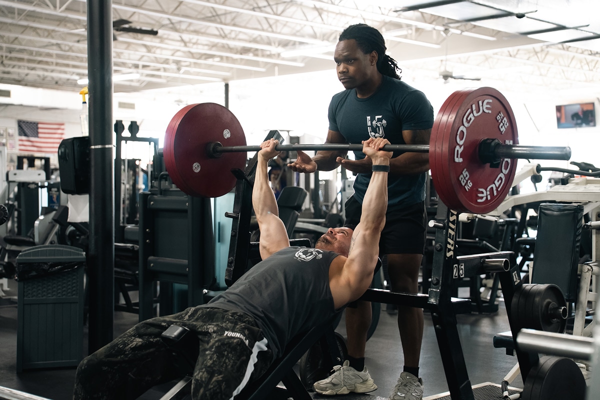 Person performing bench press with red weight plates, assisted by spotter in gym.
